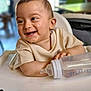 baby, bottle, high_chair, smile, happy, infant, child, face, clothing, table, arm, person, indoors, skin, head, mouth, teeth, seat, hand, background