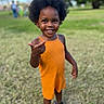 afro_hairstyle, candid, child, curly_hair, eyes, face, grass, green_background, happy, orange_outfit, outdoors, park, playful, portrait, romper, sandals, smile, standing, teeth, toddler