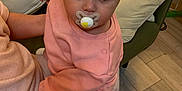 Xensia is registered to the contest to win money with this photo: baby, child, pacifier, pink_clothing, hair_clip, person, indoor, sofa, cushion, floor, wood_flooring, adult_arm, seated, looking_at_camera, cute, infant, home, cozy, portrait, closeup