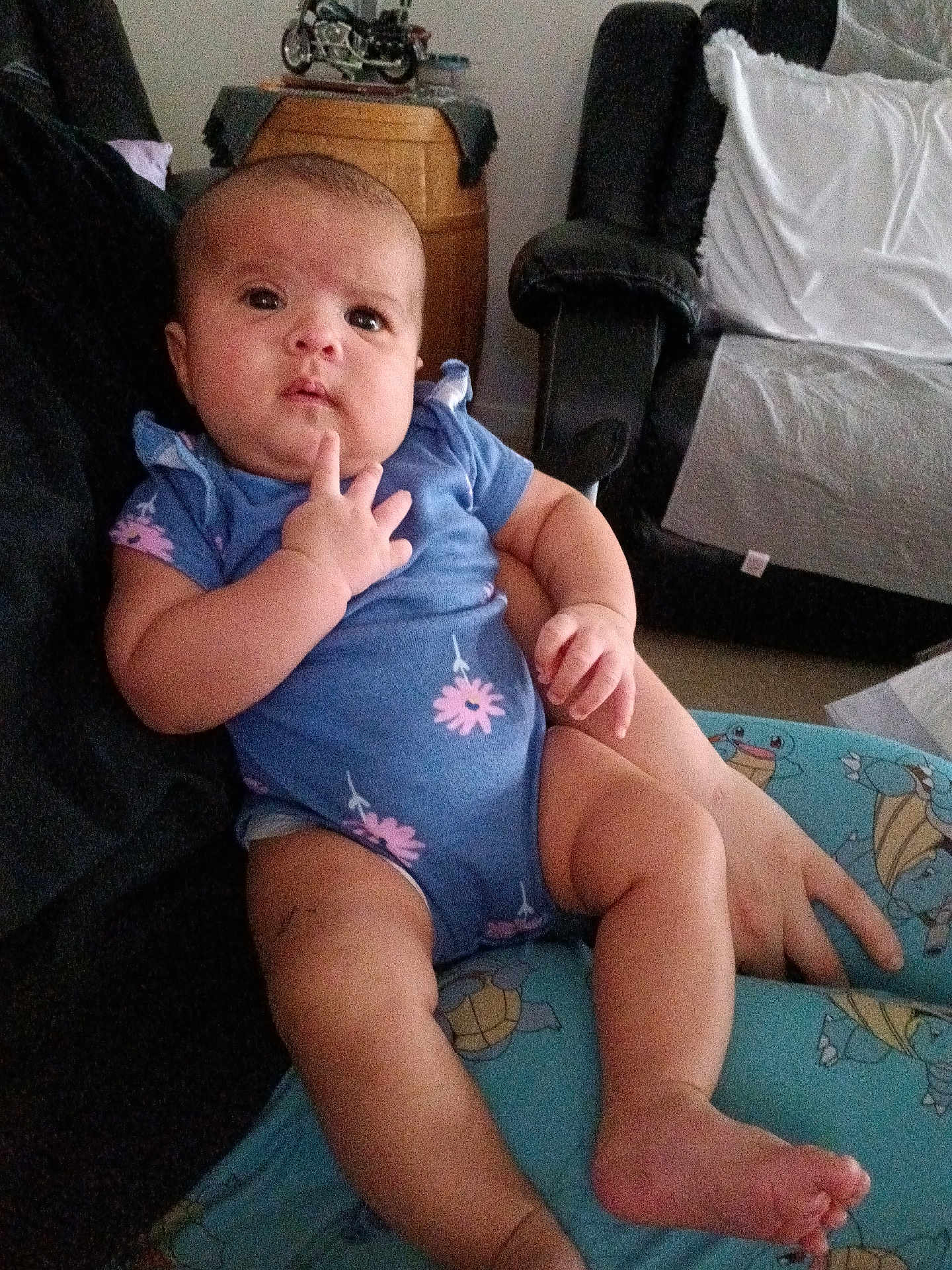 Avery is registered to the contest to win money with this photo: baby, infant, child, blue_onesie, pink_flowers, turtle_pattern, lap, hand, face, legs, barefoot, indoor, sofa, cushion, person, curious, thoughtful, skin, seated, furniture