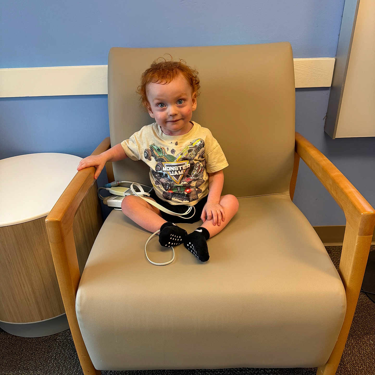 Ricky joined the competition — help win amazing prizes! toddler, child, curly_hair, sitting, armchair, beige_chair, monster_jam_tshirt, black_socks, indoor, blue_wall, round_table, wooden_armrest, carpeted_floor, smiling, young_child, cute, person, casual_clothing, curious_expression, small_furniture