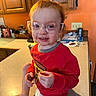 Ricky is registered to the contest to win money with this photo: toddler, child, red_hair, glasses, sweatshirt, pizza_graphic, kitchen, countertop, smiling, messy_face, sitting, indoors, cabinet, orange_wall, diaper, barefoot, toy, cup, notebook, light