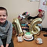 Blaise is registered to the contest to win money with this photo: child, boy, smile, birthday, balloons, cake, table, party, celebration, indoor, wall, sign, food, package, remote_control, phone, happy, casual_clothing, short_hair, sitting