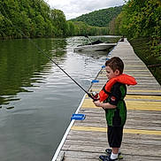 James is registered to the contest to win money with this photo: child, boy, fishing, fishing_rod, life_jacket, dock, lake, boat, water, forest, trees, hills, outdoors, nature, portrait, standing, profile, recreation, family_activity, summer