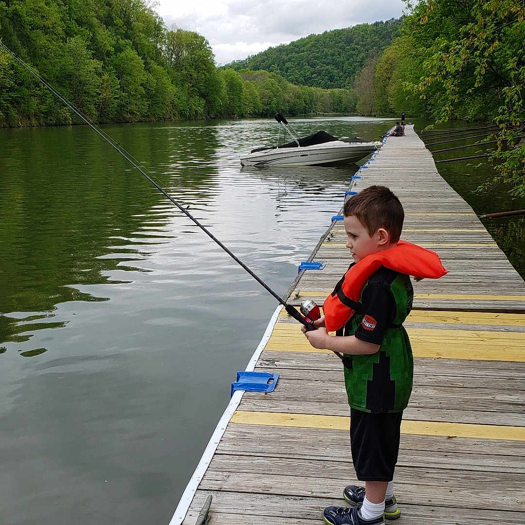James is registered to the contest to win money with this photo: child, boy, fishing, fishing_rod, life_jacket, dock, lake, boat, water, forest, trees, hills, outdoors, nature, portrait, standing, profile, recreation, family_activity, summer