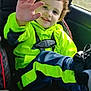 child, toddler, car_seat, bright_clothing, neon_green, navy_blue, curly_hair, smiling, waving_hand, seatbelt, car_interior, window, footwear, happy, portrait, casual_wear, young_child, safety, daylight, travel