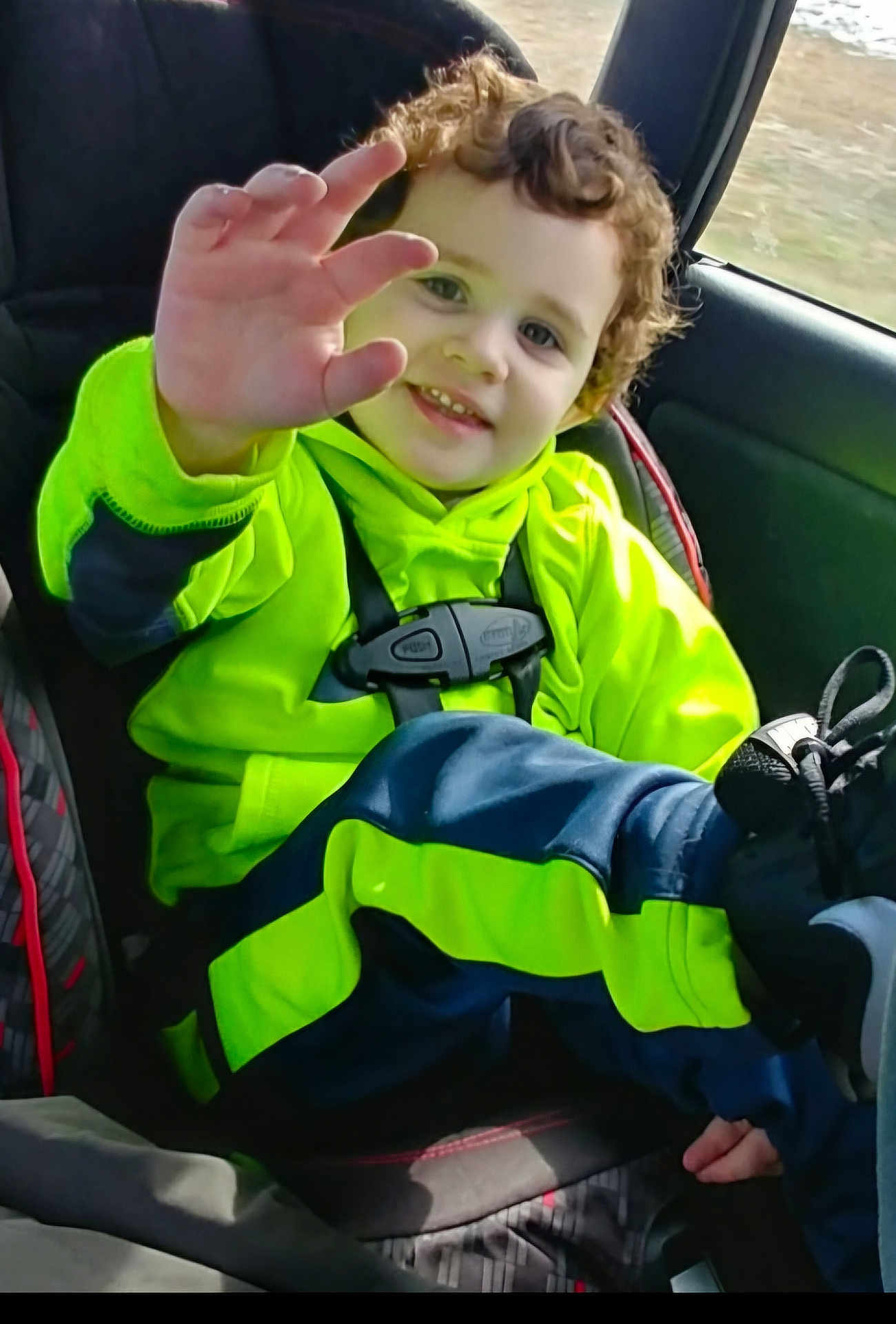 James is registered to the contest to win money with this photo: child, toddler, car_seat, bright_clothing, neon_green, navy_blue, curly_hair, smiling, waving_hand, seatbelt, car_interior, window, footwear, happy, portrait, casual_wear, young_child, safety, daylight, travel
