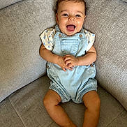Maely participe au concours pour gagner de l'argent avec cette photo : baby, child, smile, happy, romper, clothing, curly_hair, couch, furniture, indoor, person, infant, cute, skin, legs, hands, face, sitting, joyful, relaxed