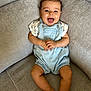 baby, child, smile, happy, romper, clothing, curly_hair, couch, furniture, indoor, person, infant, cute, skin, legs, hands, face, sitting, joyful, relaxed