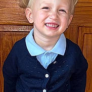 Gabriella joined the competition — help win amazing prizes! child, toddler, smiling, blonde_hair, pigtails, cardigan, blue_shirt, wooden_background, portrait, indoor, happy, cute, person, face, eyes, buttoned_clothing, standing, light_skin, young_child, casual_clothing