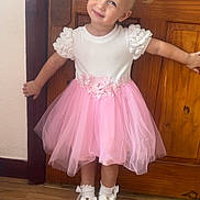 Gabriella is registered to the contest to win money with this photo: blonde_hair, bows, child, cute, fashion, floor, girl, happy, indoors, pigtails, pink_skirt, pose, smile, socks, standing, toddler, tulle_skirt, white_shoes, white_top, wooden_door