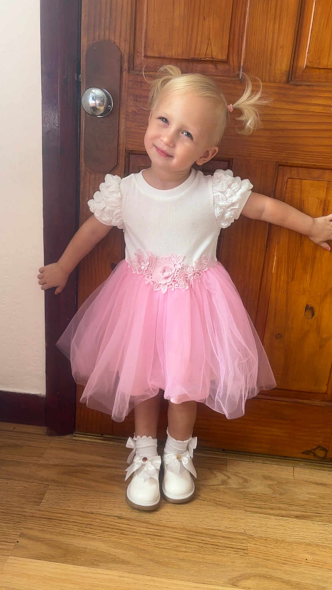 Gabriella is registered to the contest to win money with this photo: blonde_hair, bows, child, cute, fashion, floor, girl, happy, indoors, pigtails, pink_skirt, pose, smile, socks, standing, toddler, tulle_skirt, white_shoes, white_top, wooden_door