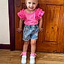 blonde_hair, casual_clothing, child, cute, denim_shorts, door_handle, flooring, girl, home, indoor, pigtails, pink_socks, pink_top, portrait, smiling, standing, toddler, white_sneakers, wooden_door, wooden_floor