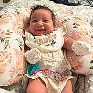 Jazlynn is registered to the contest to win money with this photo: baby, smiling, bib, floral_pillow, pacifier, bedroom, blanket, mittens, clothing, infant, cute, happy, person, lying_down, soft_toys, decor, fabric, comfort, child, portrait