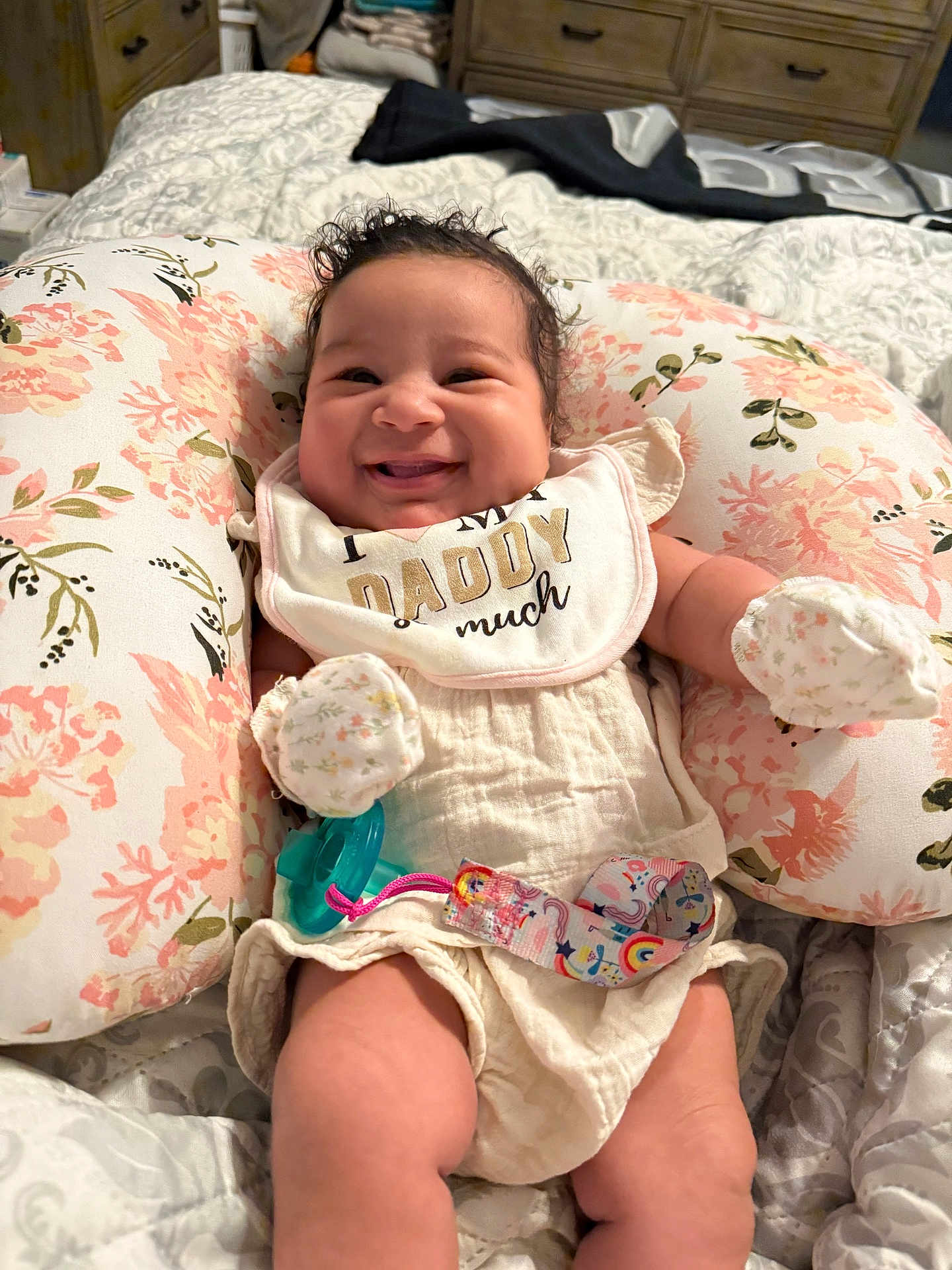 Jazlynn is registered to the contest to win money with this photo: baby, smiling, bib, floral_pillow, pacifier, bedroom, blanket, mittens, clothing, infant, cute, happy, person, lying_down, soft_toys, decor, fabric, comfort, child, portrait