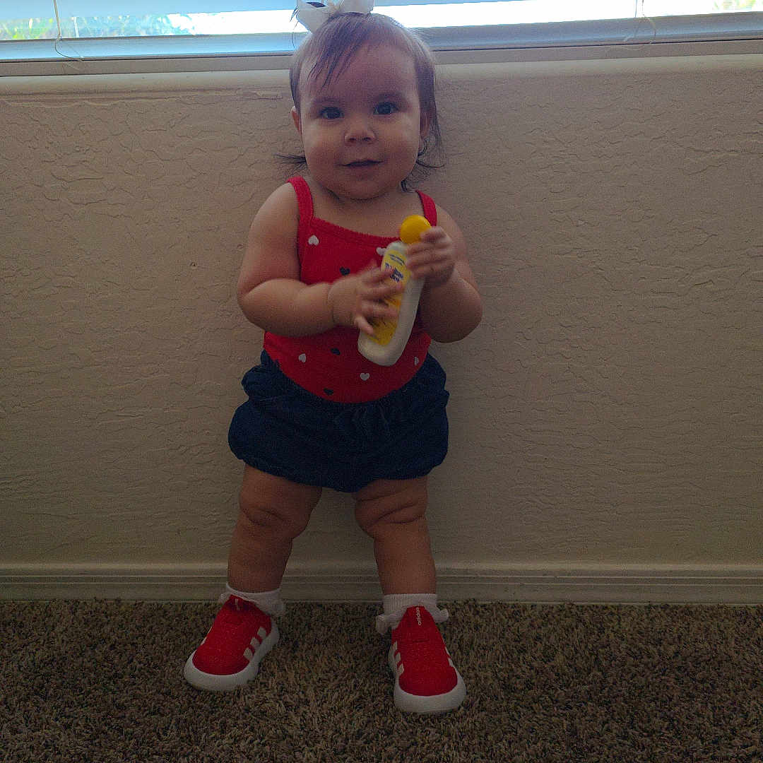 Khali is registered to the contest to win money with this photo: toddler, child, standing, carpet, red_shoes, red_top, navy_shorts, white_bow, window, blinds, bottle, indoor, wall, holding_object, infant, cute, smiling, person, young_child, hair_bow