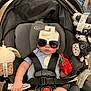 baby, car_seat, sunglasses, toy, infant, child, grape_toy, seatbelt, clothing, blue_onesie, black_straps, indoor, store, shopping, cute, expressionless, plush_toy, graco, sitting, childcare