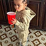 toddler, child, beige_tracksuit, hoodie, sneakers, red_cup, yellow_straw, floor_tiles, wooden_cabinet, indoor, drinking, standing, small_bottle, focused, cute, person, clothing, young_child, hair, footwear