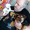 man, dog, puppy, couch, blanket, indoor, pet, holding, casual_clothing, tshirt, portrait, relaxed, companion, home, seated, love, animal, face, human, friendship