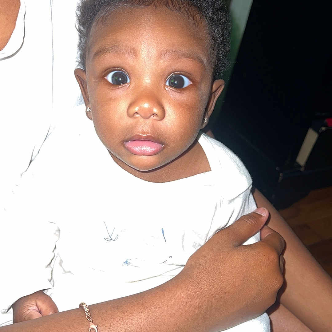 Kali joined the competition — help win amazing prizes! adult_hand, baby, bracelet, candid, child, closeup, clothing, curious, curly_hair, expression, face, holding, human, indoor, person, portrait, sitting, skin, white_clothing, wide_eyes