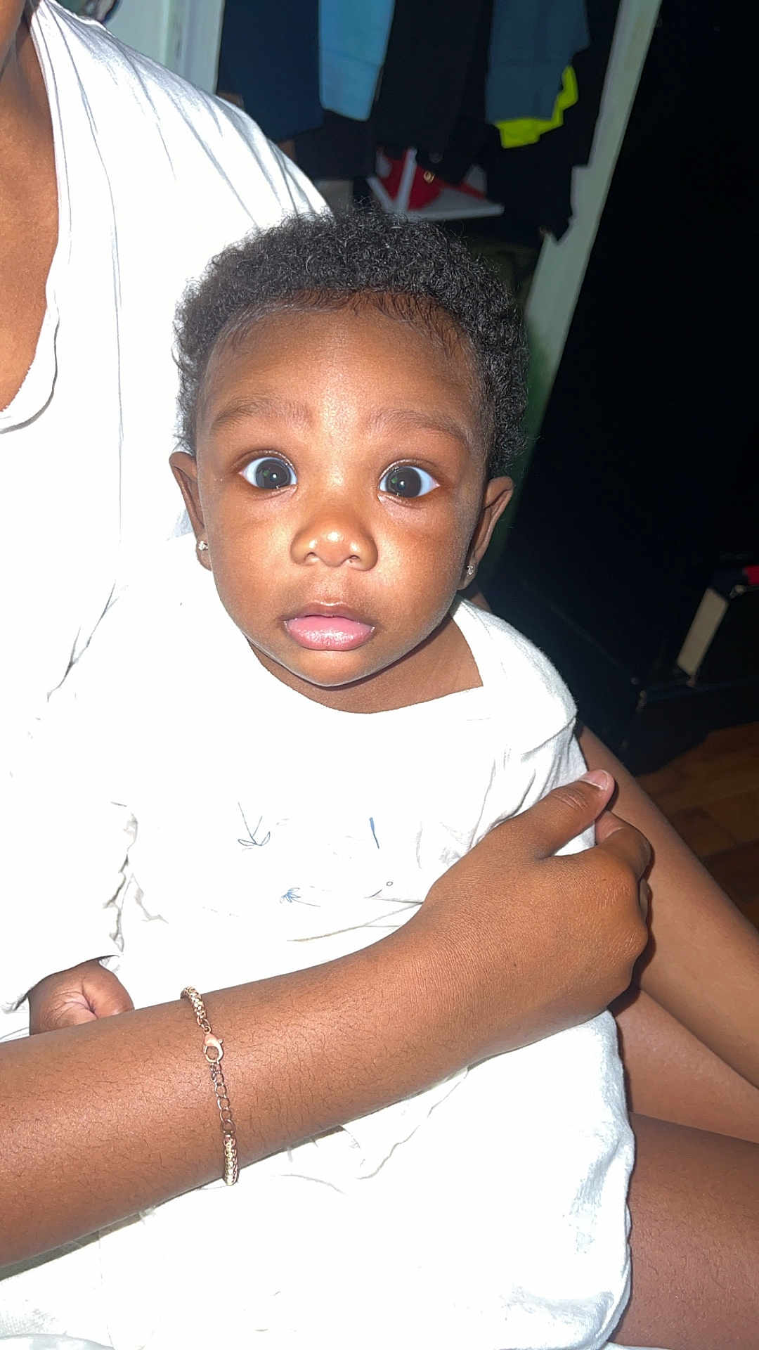 Kali joined the competition — help win amazing prizes! baby, child, curly_hair, wide_eyes, white_clothing, bracelet, adult_hand, indoor, person, holding, expression, curious, skin, face, portrait, closeup, human, sitting, clothing, candid