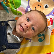 Benjamin is registered to the contest to win money with this photo: baby, infant, smiling, play_mat, colorful, cartoon, animal_print, clothing, buttoned_shirt, blue_overalls, face, head, hand, eyes, happy, cute, indoor, texture, carpet, fisherprice