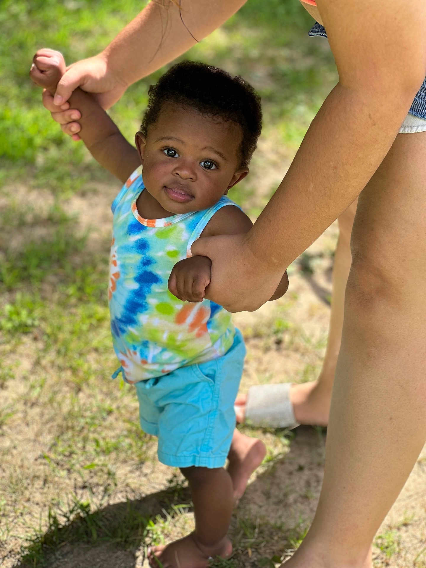 Jayden is registered to the contest to win money with this photo: toddler, child, adult, hands, grass, outdoor, sunlight, shorts, tank_top, barefoot, skin, person, support, walking, nature, summer, curly_hair, smile, daylight, holding_hands