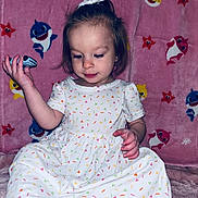 Evelyn joined the competition — help win amazing prizes! toddler, child, dress, white_dress, colorful_pattern, pink_blanket, cartoon_sharks, cartoon_starfish, indoor, sitting, cute, small_object, curious, young_child, person, portrait, soft_lighting, background_pattern, hair_bow, playful