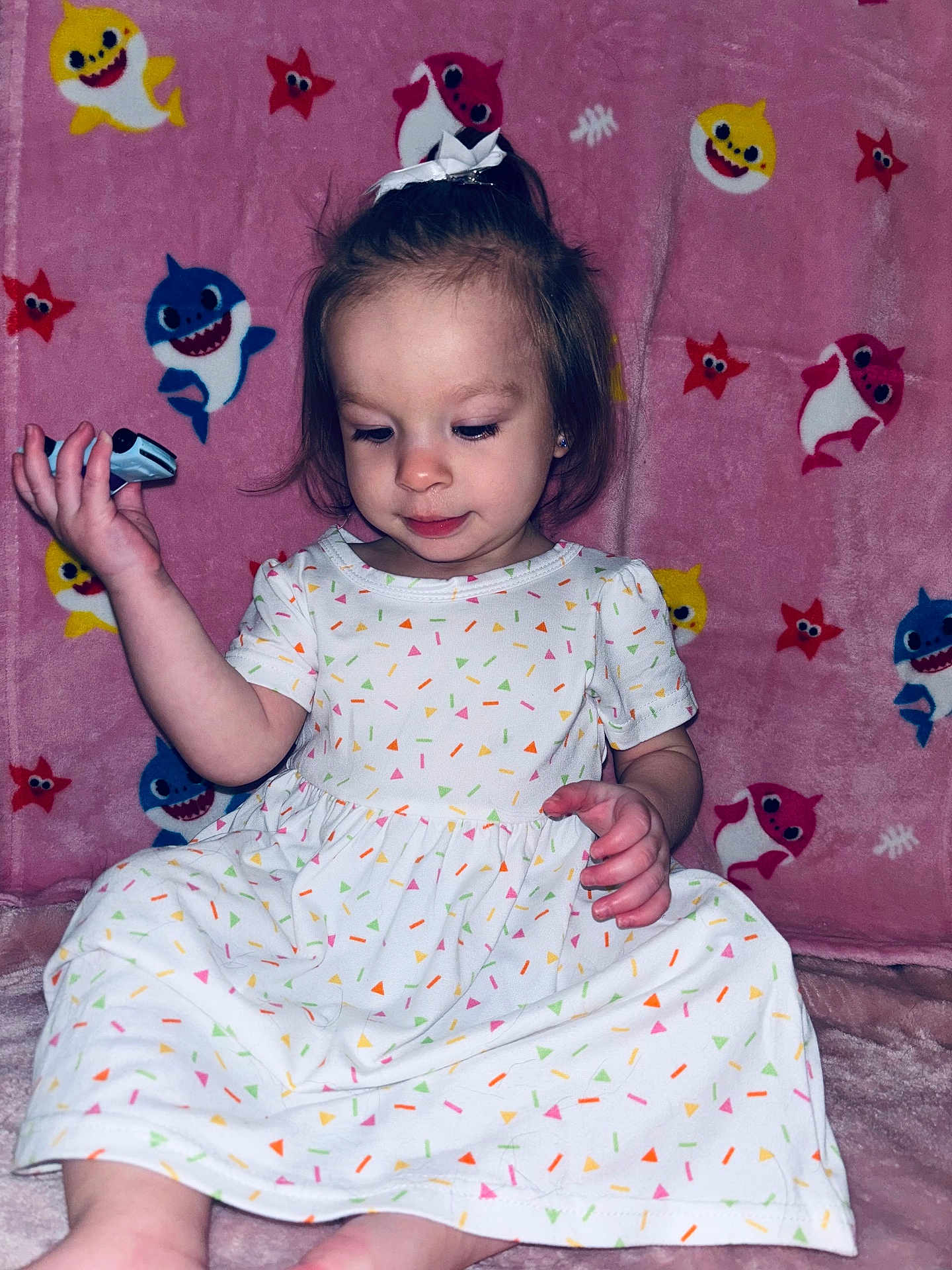 Evelyn joined the competition — help win amazing prizes! toddler, child, dress, white_dress, colorful_pattern, pink_blanket, cartoon_sharks, cartoon_starfish, indoor, sitting, cute, small_object, curious, young_child, person, portrait, soft_lighting, background_pattern, hair_bow, playful