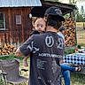 Maverick is registered to the contest to win money with this photo: baby, child, infant, face, wide_eyes, being_carried, adult, outdoor, cabin, rustic, firewood, woodpile, picnic, folding_chair, table, checkered_tablecloth, trees, grass, sunset, portrait