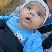 Taron-Jaques is registered to the contest to win money with this photo: baby, child, face, blue_sweater, hat, outdoor, person, expression, wide_eyes, holding, adult, brick_wall, car, nature, curious, closeup, portrait, clothing, daylight, pacifier_clip