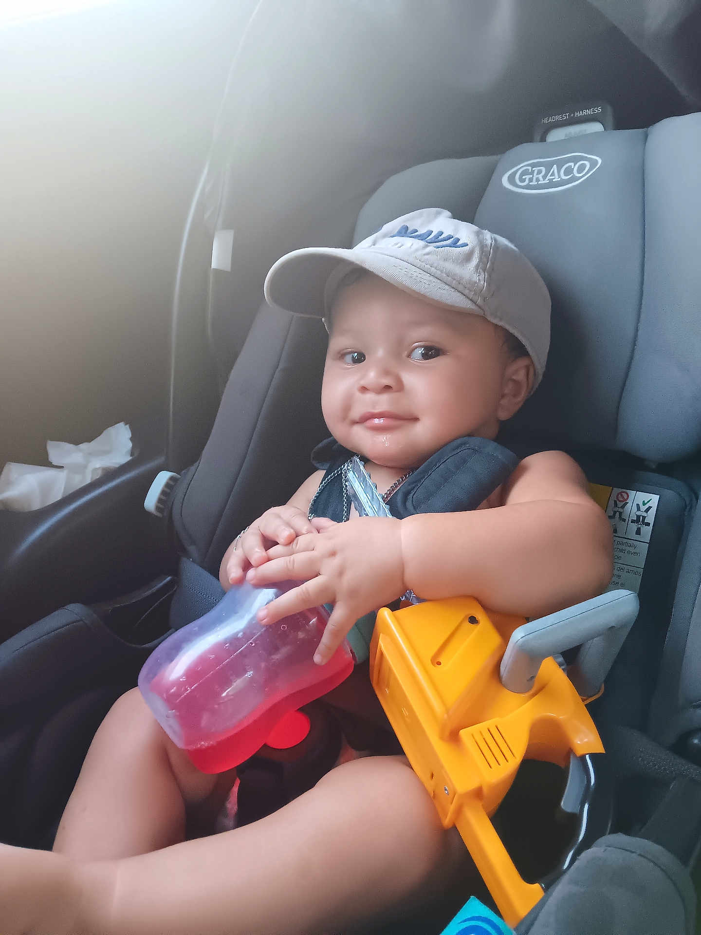Josiah is registered to the contest to win money with this photo: baby, child, car_seat, cap, toy, bottle, smile, seatbelt, infant, sitting, indoors, child_safety, graco, cute, young_child, holding, playful, car_interior, happy, person