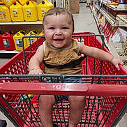 Josiah is registered to the contest to win money with this photo: baby, shopping_cart, smiling, child, store, aisle, yellow_containers, red_cart, footwear, sitting, happy, person, clothing, bracelet, floor, retail, safety_belt, toy, indoor, metal