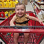 baby, shopping_cart, smiling, child, store, aisle, yellow_containers, red_cart, footwear, sitting, happy, person, clothing, bracelet, floor, retail, safety_belt, toy, indoor, metal