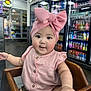 baby, child, pink_bow, high_chair, wooden_chair, cute, indoor, store, beverages, refrigerator, neon_sign, drink, sitting, clothing, buttoned_shirt, face, hand, curious, infant, portrait