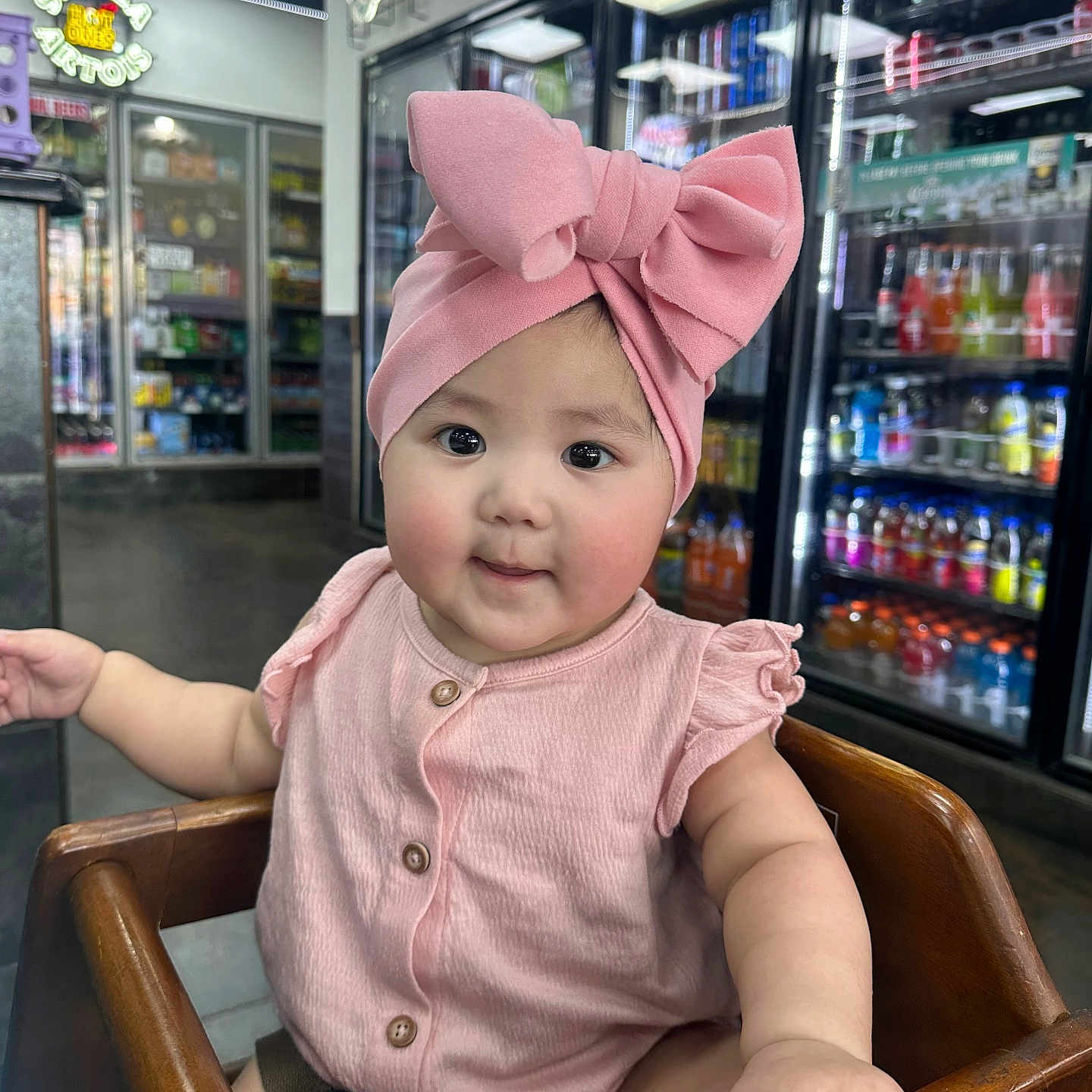 Elyesiah is registered to the contest to win money with this photo: baby, beverages, buttoned_shirt, child, clothing, curious, cute, drink, face, hand, high_chair, indoor, infant, neon_sign, pink_bow, portrait, refrigerator, sitting, store, wooden_chair