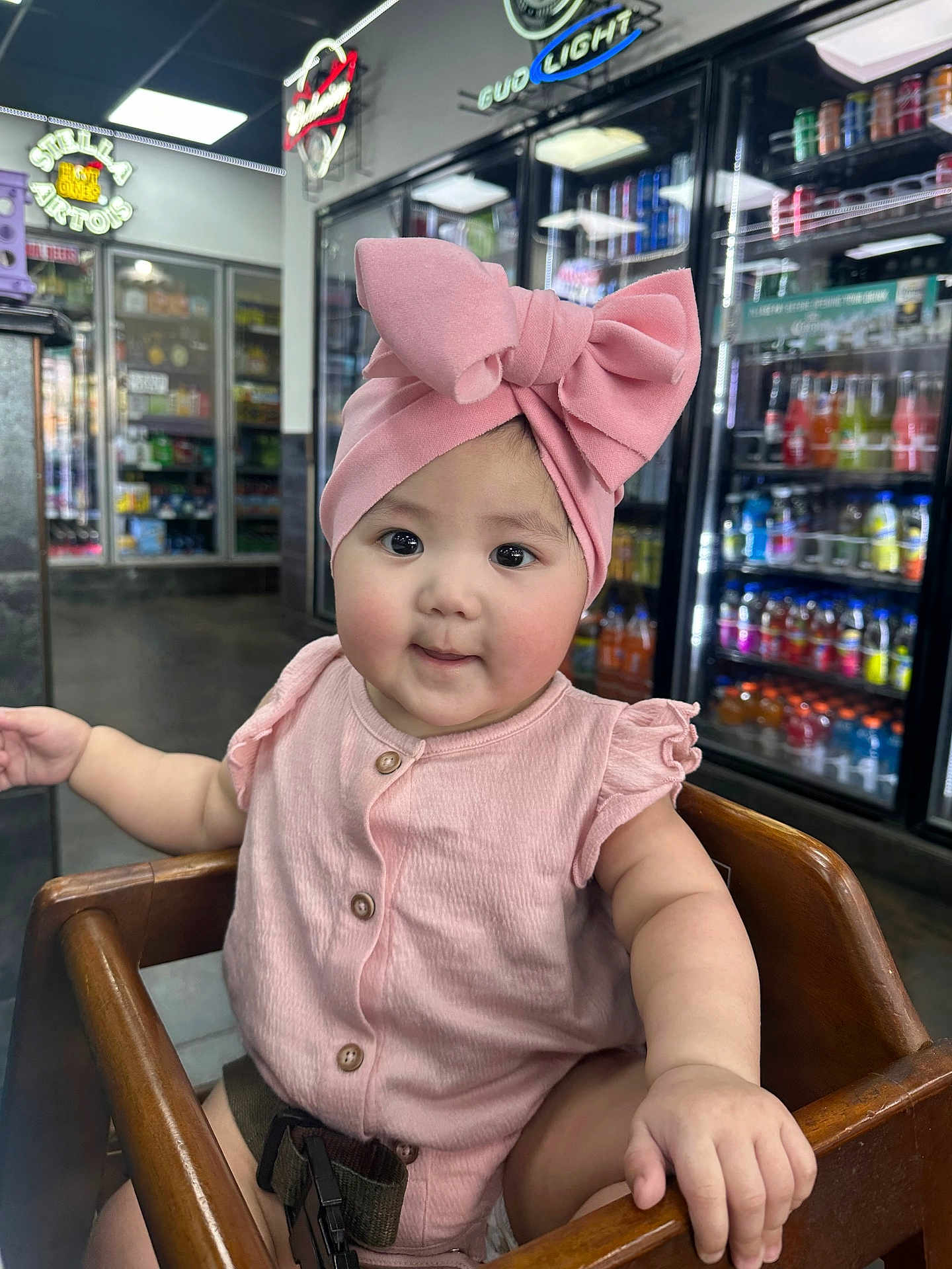 Elyesiah is registered to the contest to win money with this photo: baby, child, pink_bow, high_chair, wooden_chair, cute, indoor, store, beverages, refrigerator, neon_sign, drink, sitting, clothing, buttoned_shirt, face, hand, curious, infant, portrait