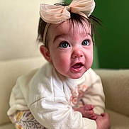 Mia is registered to the contest to win money with this photo: baby, child, headband, bow, pink_bow, sitting, clothing, white_sweater, floral_pants, chair, cream_chair, indoor, portrait, expression, cute, face, wide_eyes, person, soft_lighting, background_blur