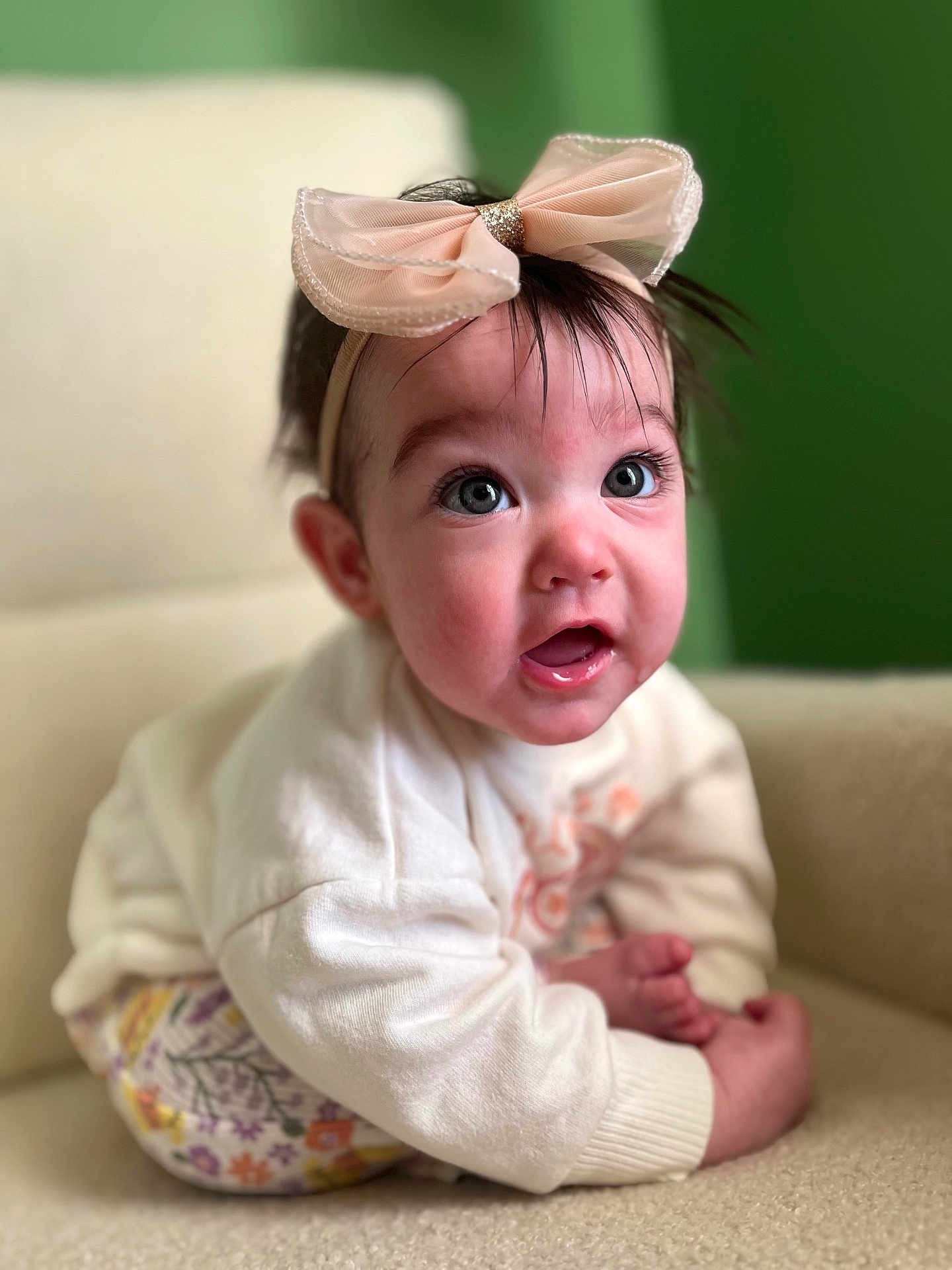 Mia is registered to the contest to win money with this photo: baby, child, headband, bow, pink_bow, sitting, clothing, white_sweater, floral_pants, chair, cream_chair, indoor, portrait, expression, cute, face, wide_eyes, person, soft_lighting, background_blur