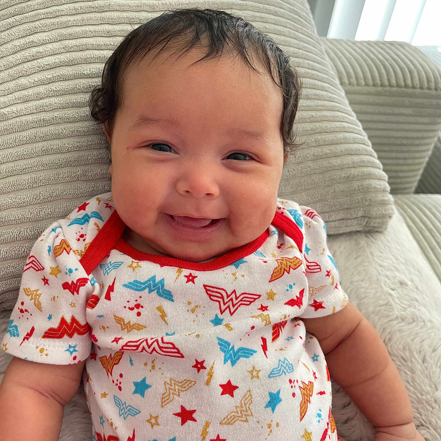Aria is registered to the contest to win money with this photo: baby, child, chubby_cheeks, clothing, couch, cute, fabric, furniture, happy, home, indoor, infant, light, onesie, person, portrait, relaxed, smiling, texture, wonder_woman