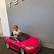 Kaarvik joined the competition — help win amazing prizes! toddler, child, toy_car, red_car, tesla, indoor, car_showroom, logo, wall, floor, carpet, person, small_vehicle, smiling, happy, cute, modern, technology, electric_car, vehicle