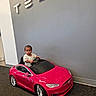 toddler, child, toy_car, red_car, tesla, indoor, car_showroom, logo, wall, floor, carpet, person, small_vehicle, smiling, happy, cute, modern, technology, electric_car, vehicle