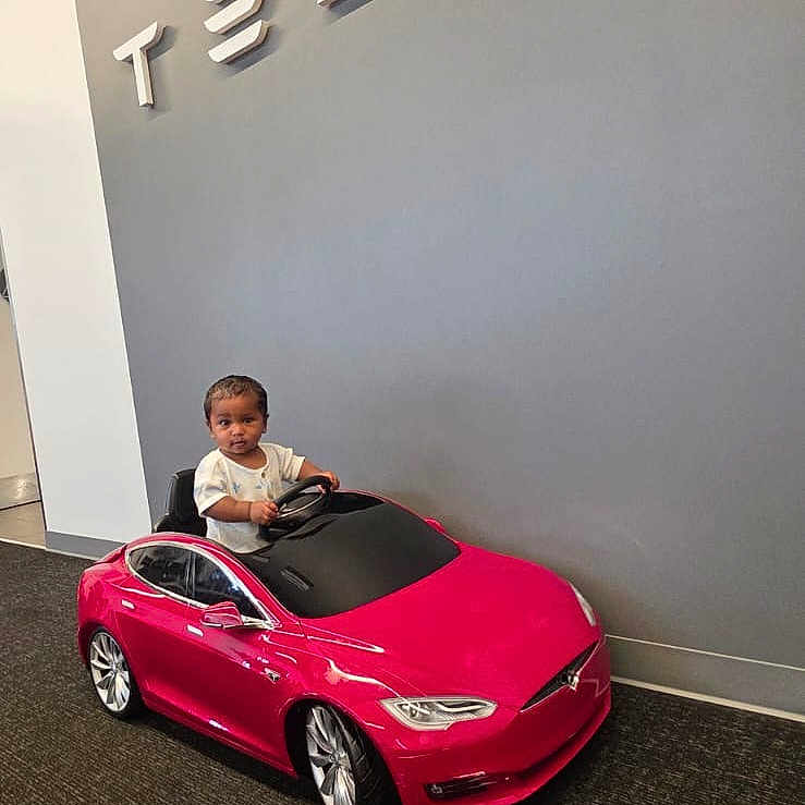 Kaarvik joined the competition — help win amazing prizes! toddler, child, toy_car, red_car, tesla, indoor, car_showroom, logo, wall, floor, carpet, person, small_vehicle, smiling, happy, cute, modern, technology, electric_car, vehicle