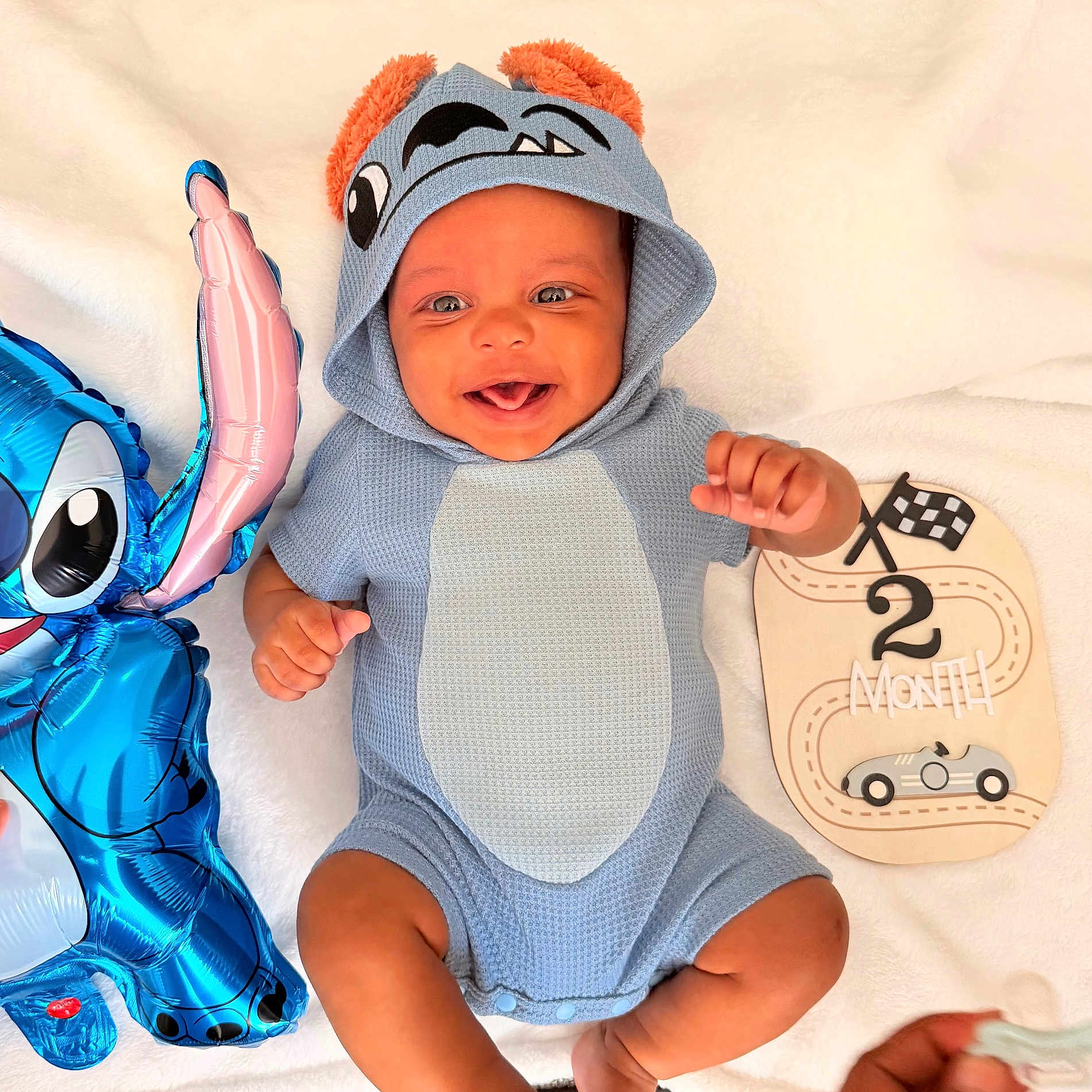 Jonas is registered to the contest to win money with this photo: baby, infant, smile, blue_onesie, hood, orange_ears, balloon, cartoon_character, milestone_board, 2_month, racing_car, soft_blanket, happy, cute, portrait, child, lying_down, hand, face, expression