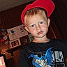 child, boy, red_cap, batman_tshirt, blue_shorts, indoor, wood_paneling, children_artwork, television, casual_pose, portrait, person, face, headwear, smiling, short_hair, blue_eyes, home_interior, young, cute
