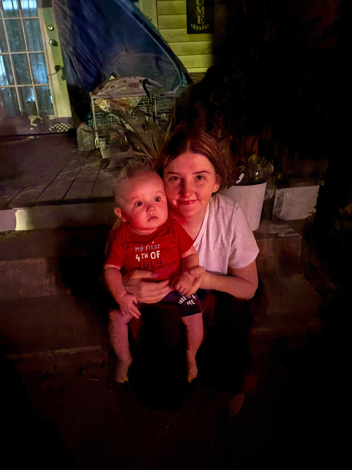 Emilio is registered to the contest to win money with this photo: baby, woman, child, porch, night, outdoor, plants, steps, red_shirt, celebration, holding, smile, face, person, home, door, window, clothing, dark, expression
