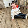 child, porch, wooden_deck, sweatshirt, pants, white_shoes, door, pet_flap, outdoor, curious_expression, small_pony_tail, casual_clothing, wooden_railing, vehicle, toy, floor, hand, sitting, daylight, person