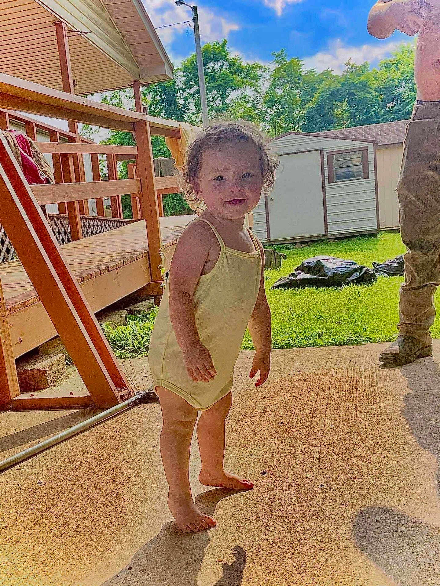 Hazel Hamby joined the competition — help win amazing prizes! toddler, child, yellow_romper, barefoot, outdoor, patio, concrete, wooden_deck, greenery, grass, shed, sunlight, shadow, adult, shirtless, summer, smiling, curly_hair, happy, family
