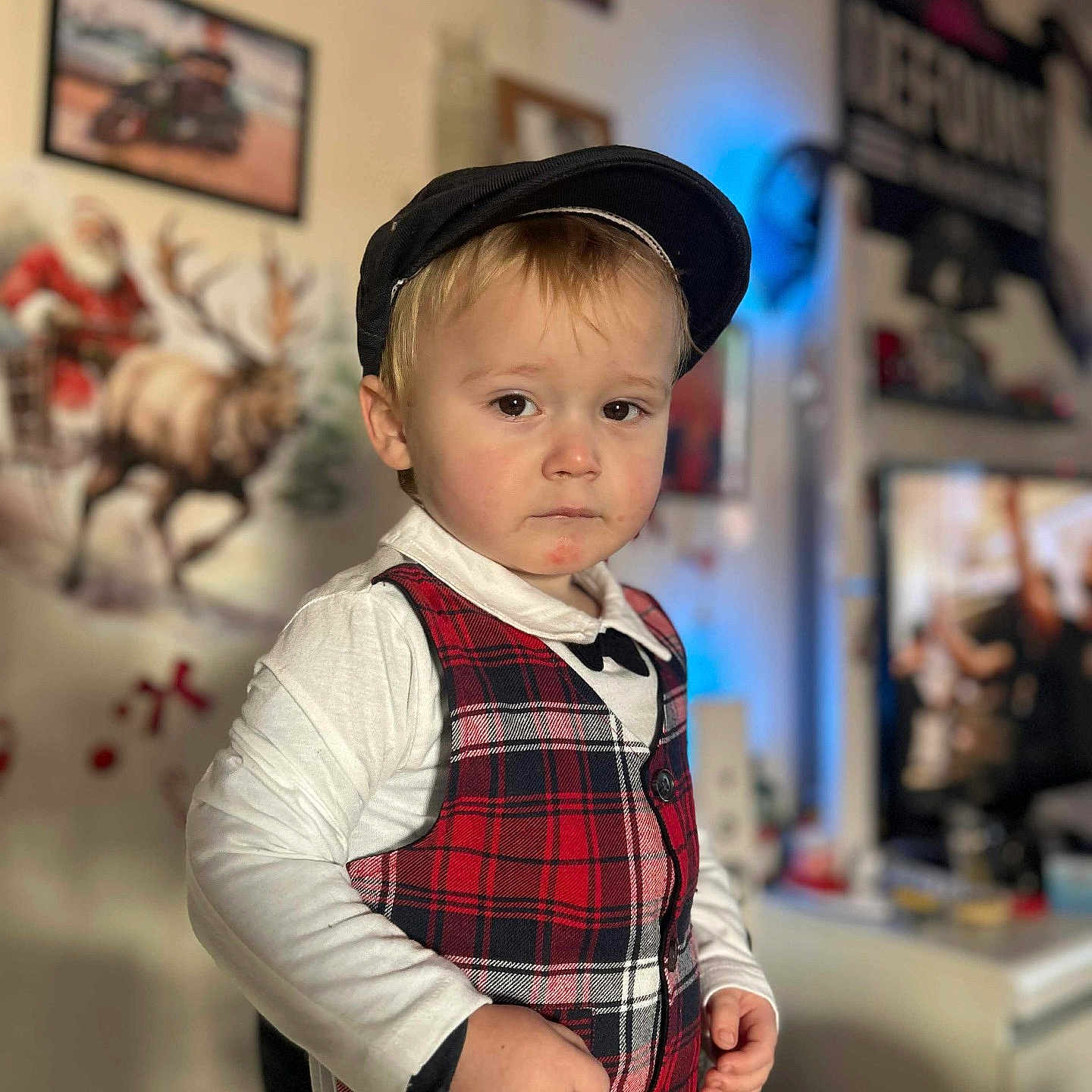 Cylian a rejoint le concours — aidez-le/la à gagner de superbes lots ! blurred_background, bow_tie, boy, cap, casual_clothing, child, cute, expression, flat_cap, framed_picture, hands, holiday_decor, indoor, living_room, moose_painting, plaid_vest, portrait, shallow_depth_of_field, toddler, tv_screen