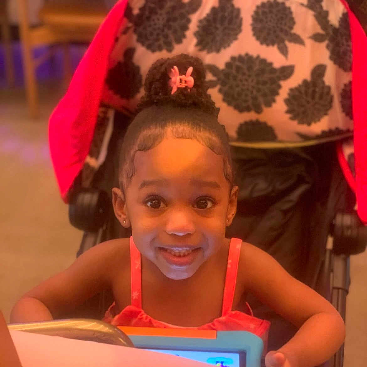 Oasis is registered to the contest to win money with this photo: child, smiling, tablet, stroller, hair_bun, pink_clip, indoor, floral_pattern, happy, young_child, person, technology, seated, bright_lighting, face, cute, playful, clothing, table, expression
